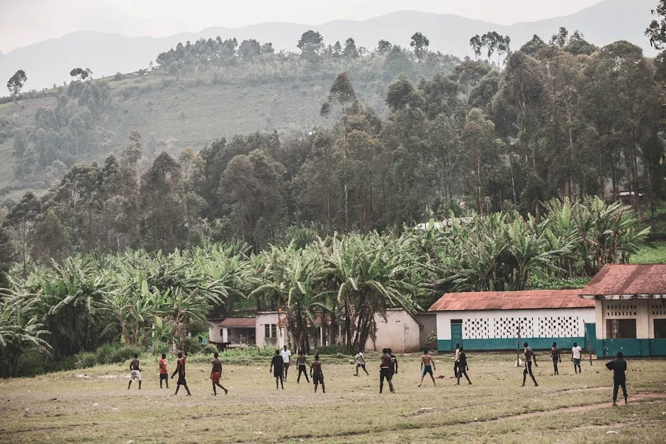 Children play soccer on a grassy field surrounded by lush African landscape and small village buildings.