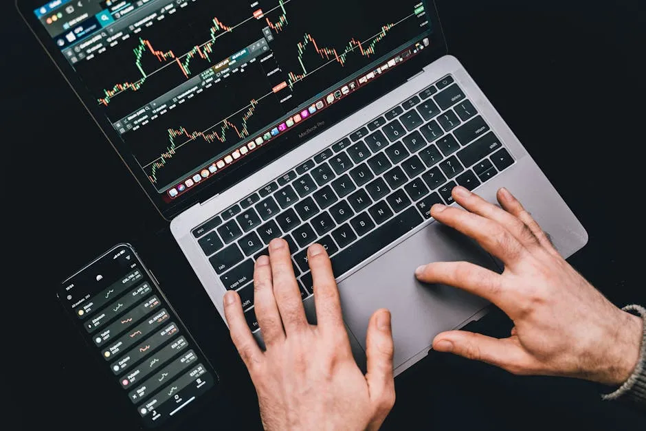 Hands typing on a laptop displaying stock market charts and a smartphone with financial apps.