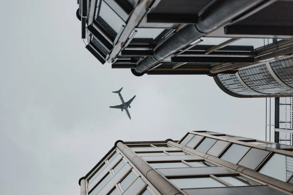 Low angle view of an airplane flying over modern skyscrapers in a cloudy sky.