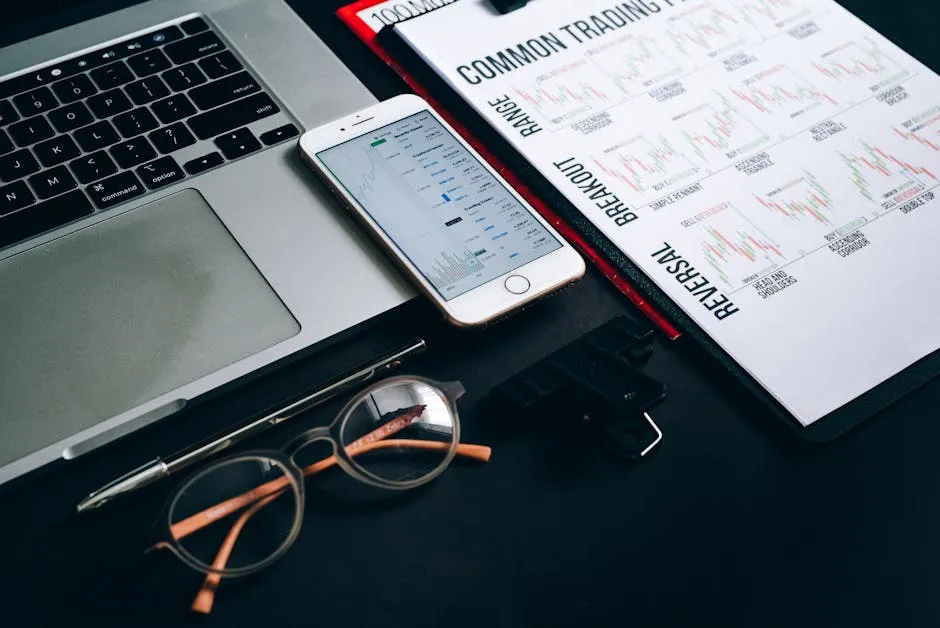 Workstation with laptop, smartphone, eyeglasses, financial charts, and pen for trading analysis.